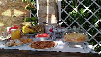 Un bufé de desayuno con diversos alimentos, entre ellos frutas, pastelería y un pastel, montado al aire libre con una valla de celosía y follaje en el fondo.