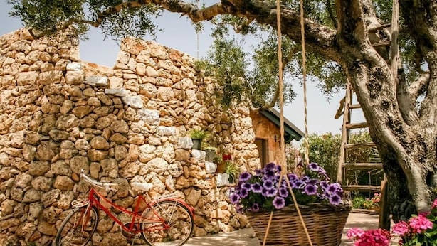 Un muro de piedra, una bicicleta roja y una cesta de flores moradas.