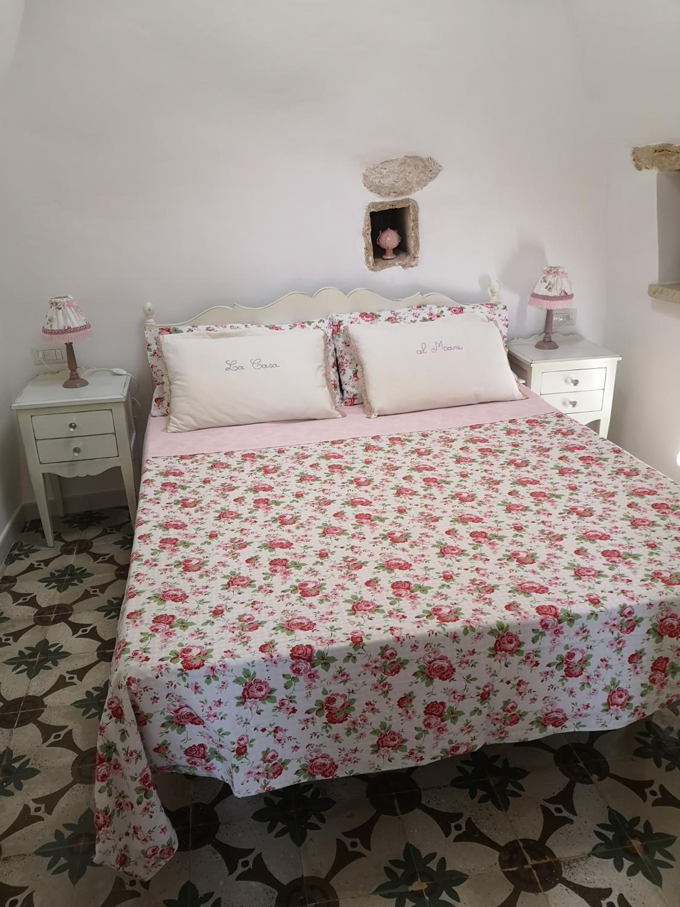 Un dormitorio con edredón estampado floral, dos almohadas con texto, una mesita de noche con lámpara y un estante fijado a la pared.