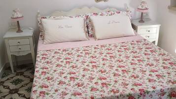 Un dormitorio con edredón estampado floral, dos almohadas con texto, una mesita de noche con lámpara y un estante fijado a la pared.