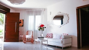Una sala de estar moderna con un sofá blanco, una mesita redonda pequeña, una silla de mimbre y una planta en maceta con flores rojas.
