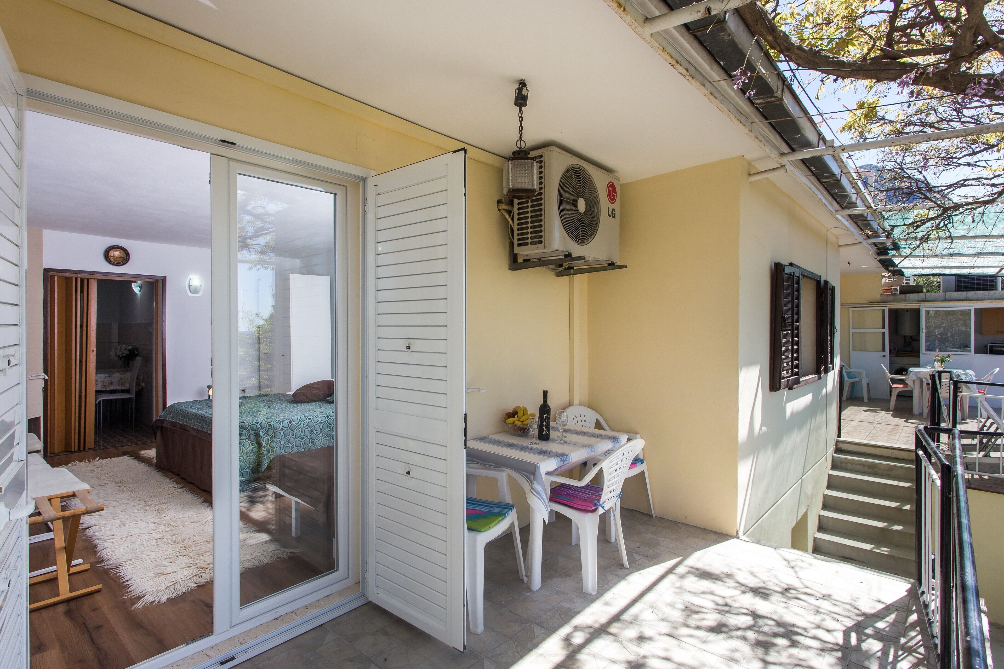 Un balcón con puerta corredera de vidrio que da a un dormitorio, una mesa con sillas y un equipo de aire acondicionado instalado en la pared.
