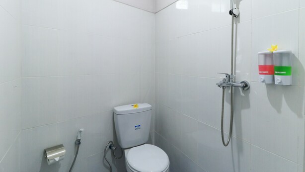 Une petite salle de bain aux murs recouverts de carreaux blancs, équipée de toilettes, d’une douchette et d’un distributeur de savon fixé au mur.