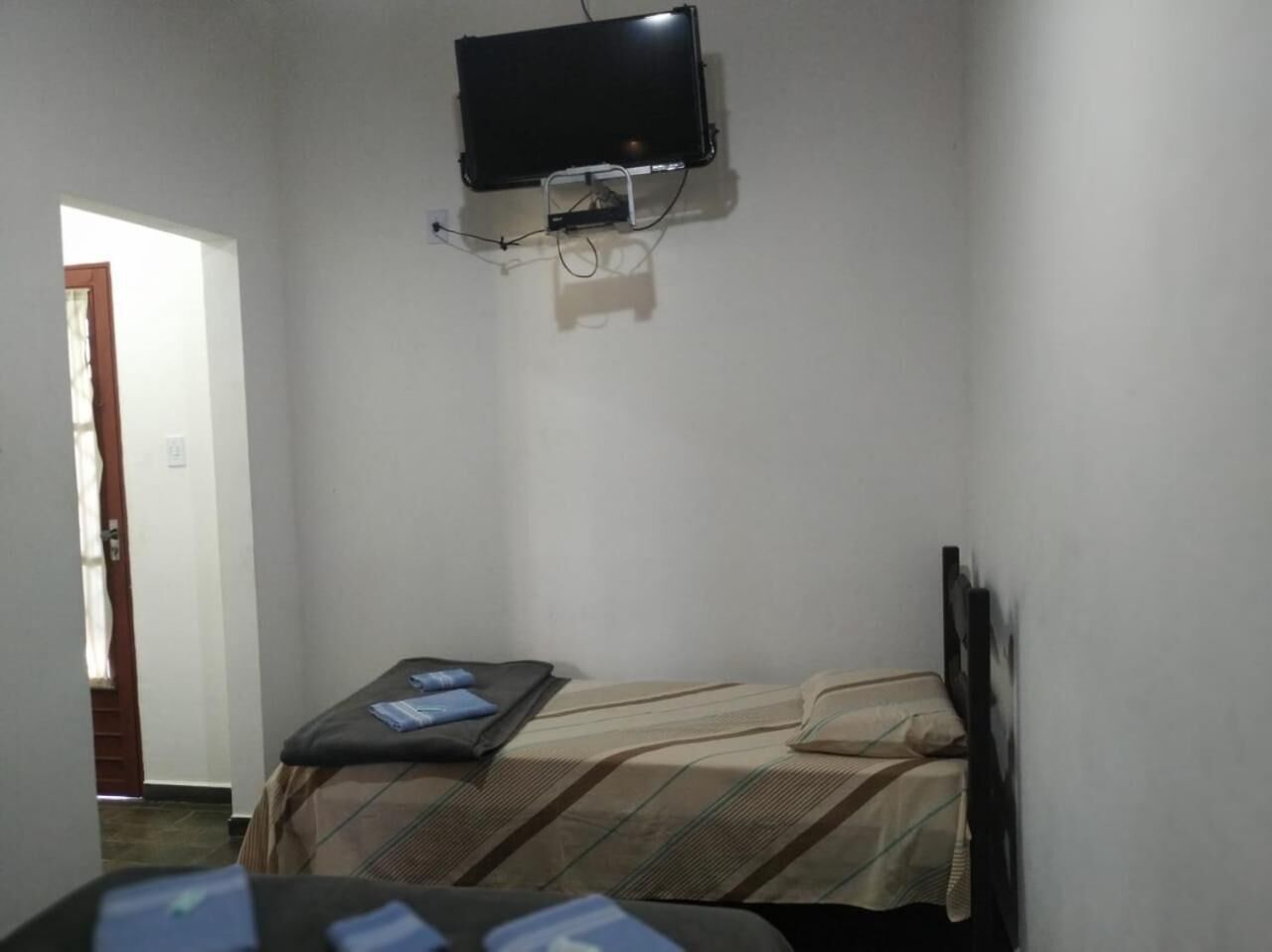 Quarto pequeno e modesto, com uma cama, uma escrivaninha e uma televisão na parede.