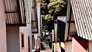 Un estrecho callejón entre edificios con techos de lámina corrugada, que conduce a un cartel con indicaciones hacia 'Churrasco', 'Salero-Juegos' y 'Piscina'.