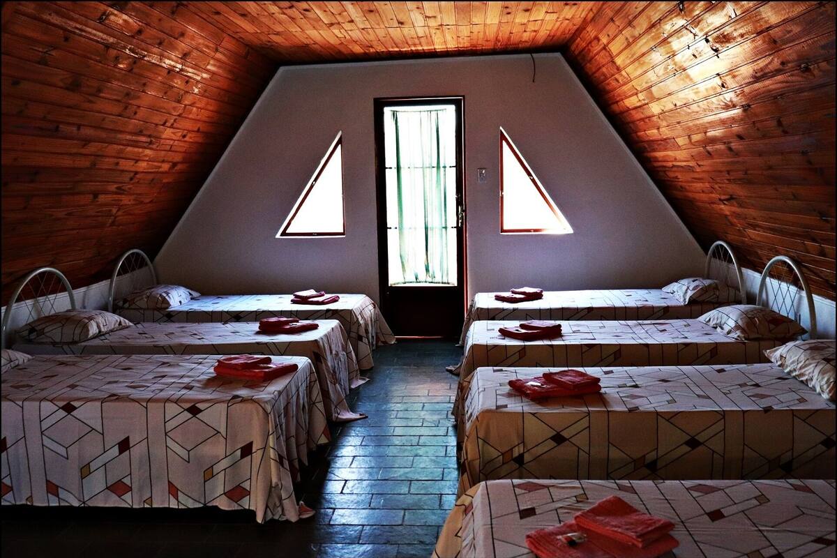 Un dormitorio abuhardillado triangular con cuatro camas dispuestas en fila, cada una con una colcha estampada y una toalla roja.
