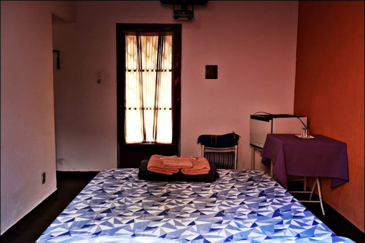 Una habitación de hotel con una colcha de diseño geométrico, una pila de toallas ordenadas, una ventana con cortinas, una mesita con mantel morado y un altavoz sobre un soporte.