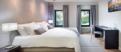 A modern bedroom with a large bed, a desk, and a view of greenery outside.