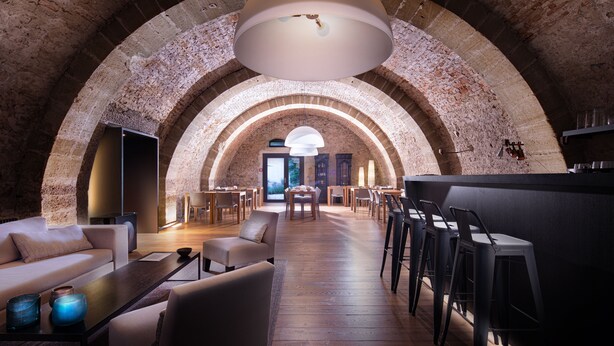 Um espaço amplo, com teto em arco, mesa central e cadeiras, área de bar com banquetas e uma luminária redonda grande.