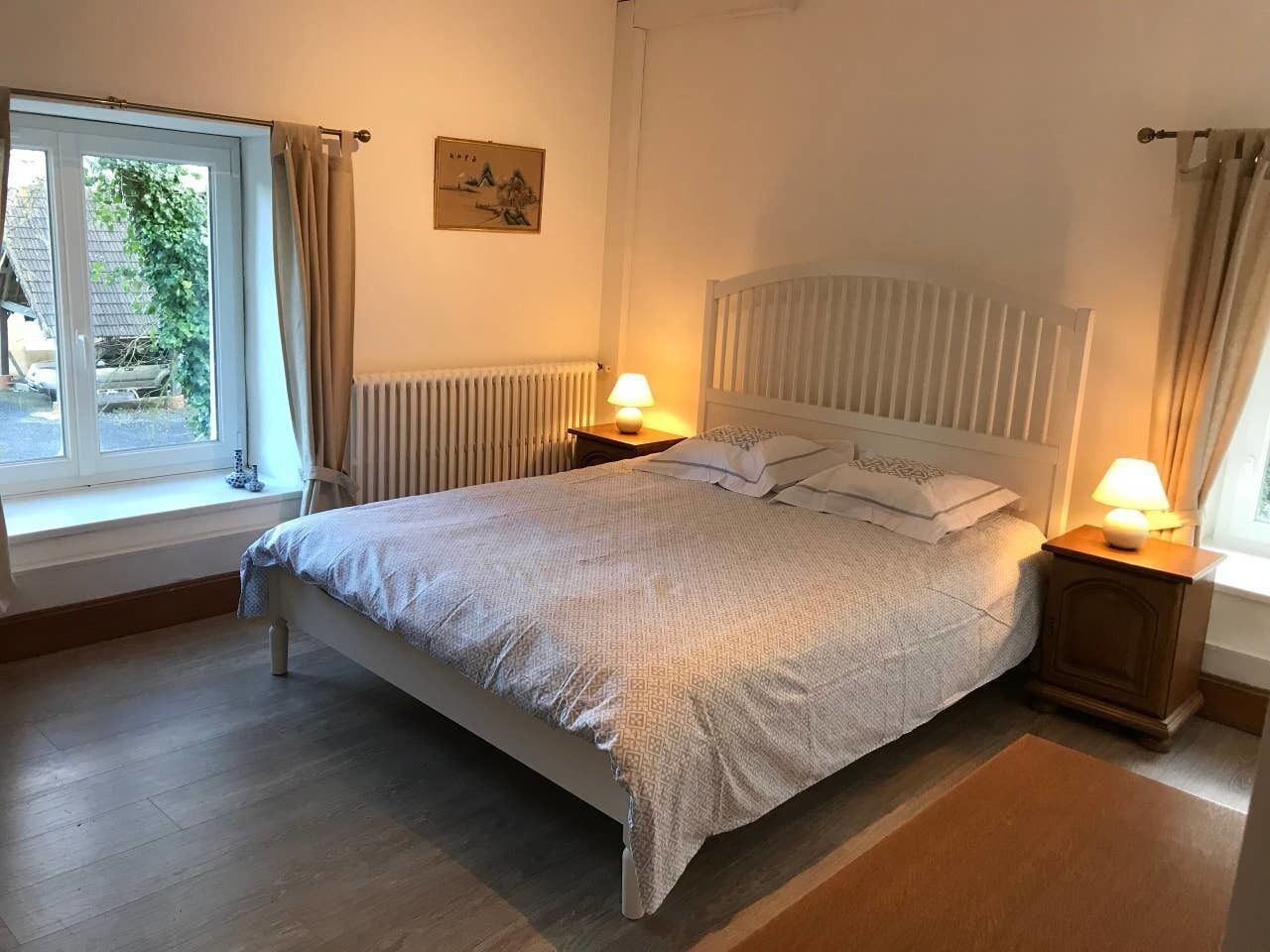 Un dormitorio con una cama grande, dos mesitas de noche con lámparas, un radiador y una ventana con cortinas.