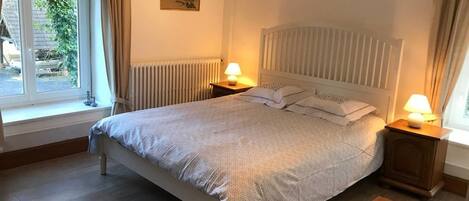 Un dormitorio con una cama grande, dos mesitas de noche con lámparas, un radiador y una ventana con cortinas.