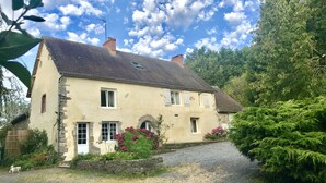 A two-story stone house with a gravel driveway, surrounded by greenery and flowering plants.