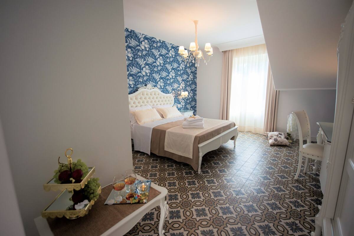 Camera da letto con un grande letto, un lampadario, pavimento piastrellato a motivi e un comodino con un vassoio di frutta e un bicchiere.