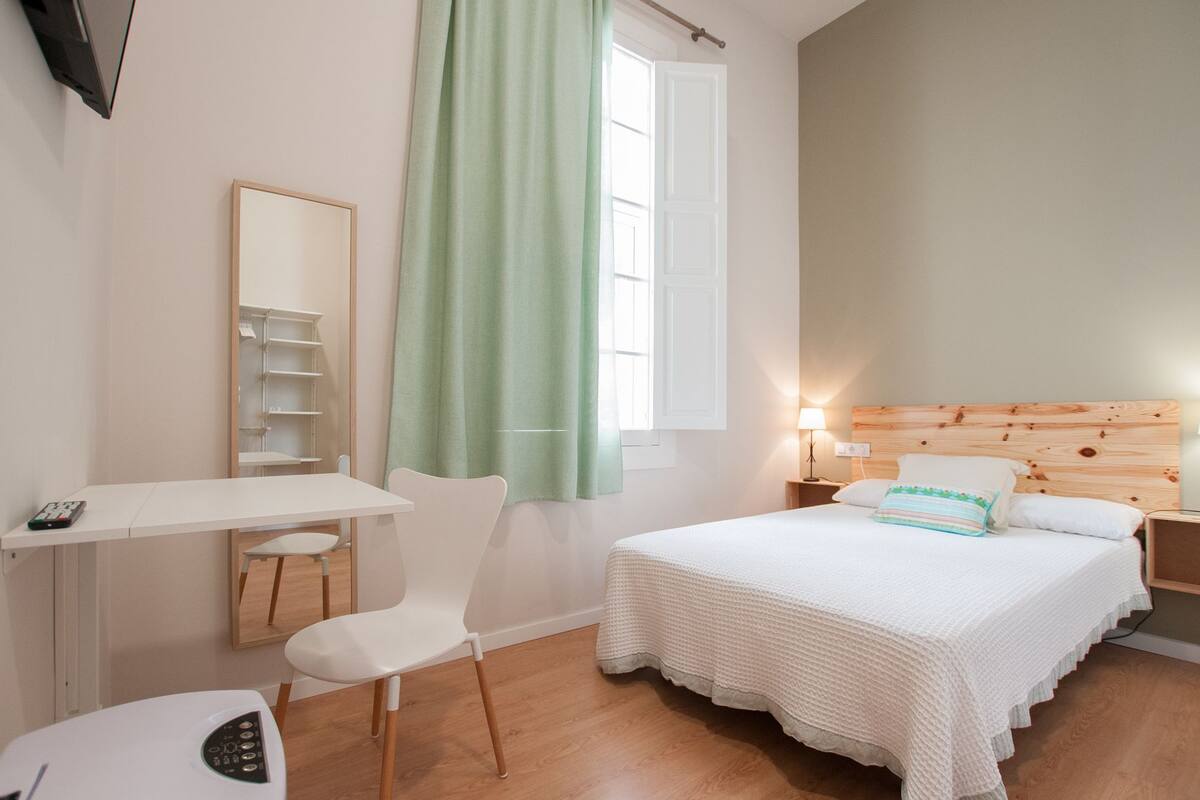 Un dormitorio con cama, escritorio, silla, espejo, ventana con cortinas y lámpara de noche.