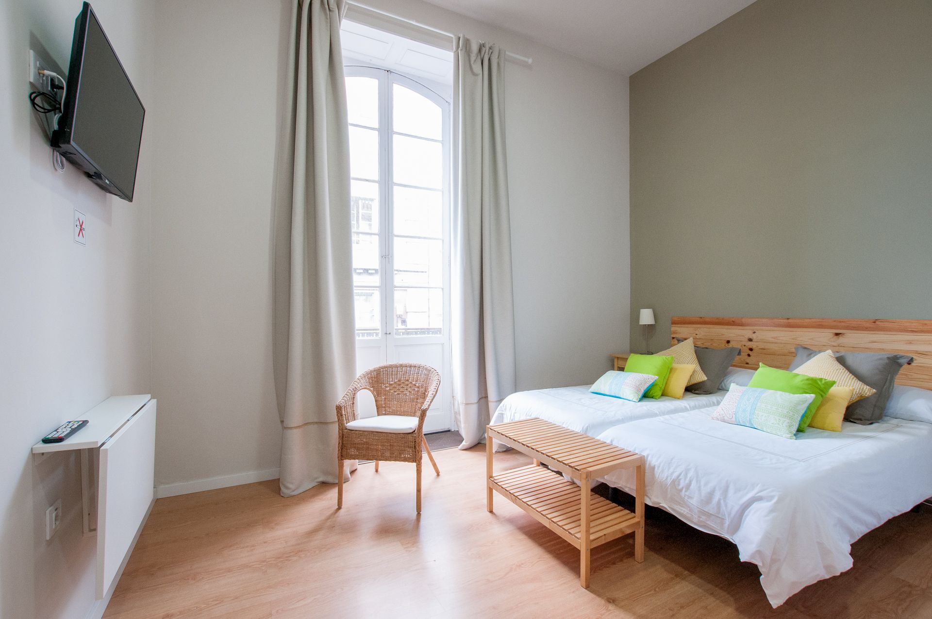 A bedroom with a bed, a chair, a small table, a TV mounted on the wall, and a window with curtains.