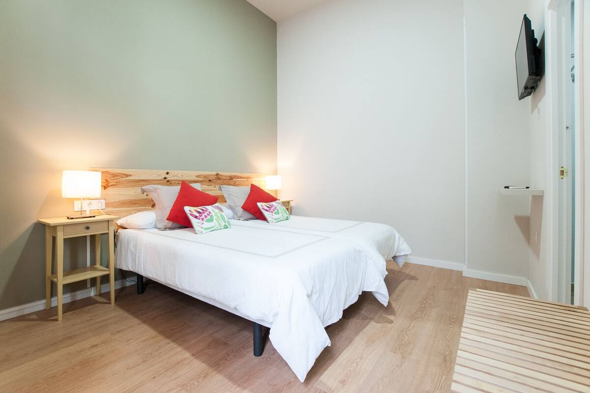 Un dormitorio ordenado con cabecera de madera, una mesita de noche con lámpara y una cama con ropa de cama blanca y almohadas rojas.