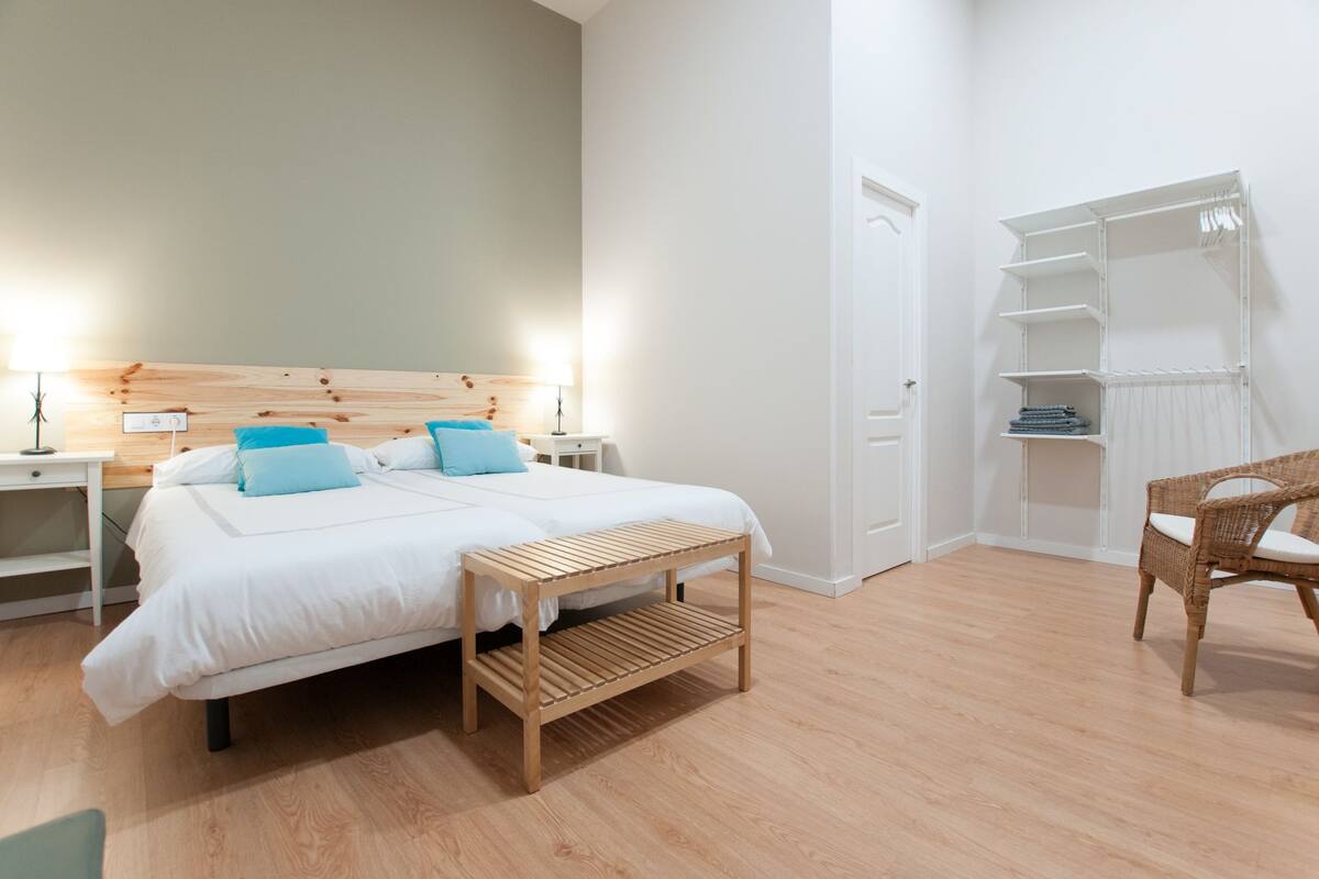 Un dormitorio con cama doble, mesitas de noche, un banco de madera, una silla de mimbre y un armario blanco con estantes.