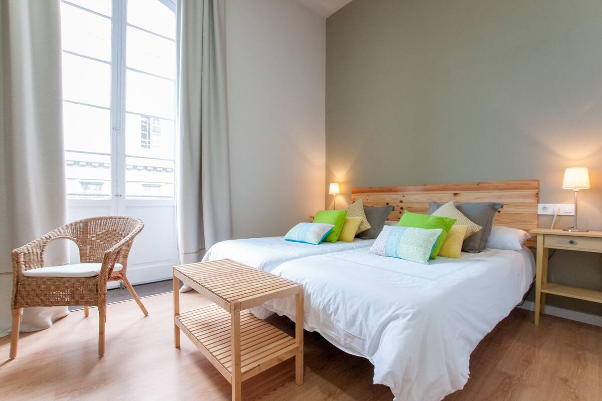 Habitación de hotel con dos camas, una silla de mimbre, una mesita de madera y una mesita de noche con lámpara.