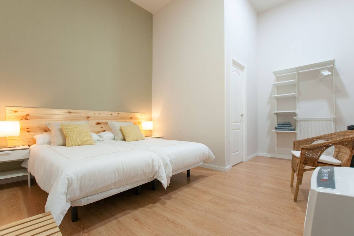 Un dormitorio con cabecera de madera, cama con ropa de cama blanca, dos lámparas de noche, una silla de mimbre y un armario blanco con estantes.