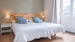 Ein Doppelbett mit weißen Leinentüchern, zwei Nachttische mit Lampen, ein hölzernes Kopfteil und ein großes Fenster mit Vorhängen.