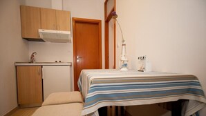 Une petite pièce, de taille modeste, comprenant un coin cuisine, un lit recouvert d’une couverture à rayures et une table de chevet avec une lampe.