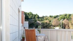 Une chaise et une table en bois sur un balcon doté d’un sol carrelé, entourés d’une balustrade blanche, avec en arrière-plan une vue sur des arbres et des bâtiments.