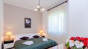 A hotel room with a bed, two bedside tables with lamps, a nightstand, a wooden table, and a vase with flowers. - Apartments Nicol - Two-Bedroom Apartment with Balcony and Sea View (Dubrovnik)