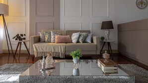 A living room with a beige sofa, decorative pillows, a glass coffee table with books and a plant, a side table with a lamp, and a small plant on a stand. - Hotel Canto Do Chafariz (Diamantina)