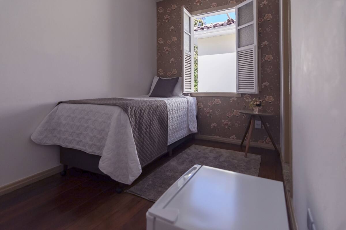 A small bedroom with a bed, a window with shutters, a small table, and a rug.