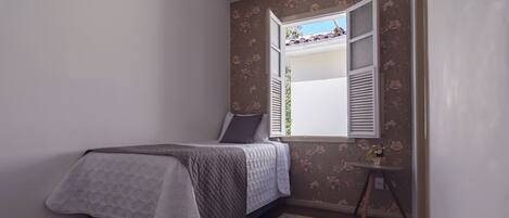 Um quarto pequeno com uma cama, uma janela com persianas, uma mesa e um tapete.