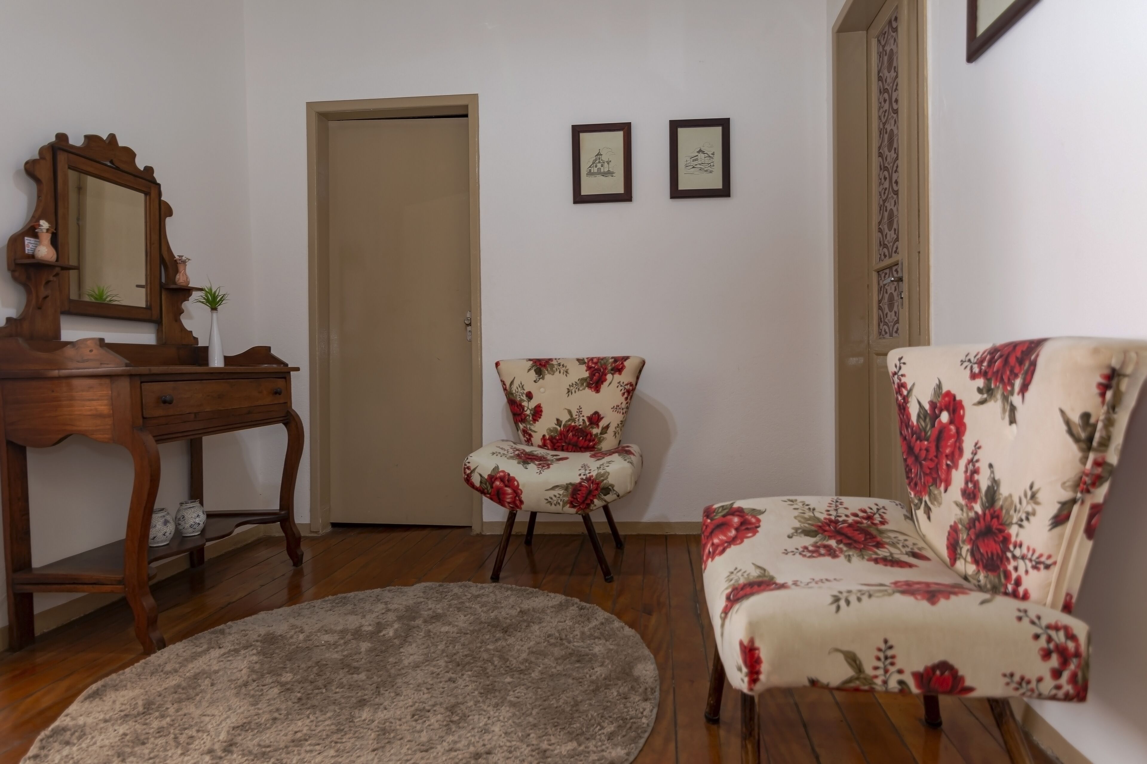 A room with a wooden desk, two chairs with floral upholstery, a round rug, and a door.