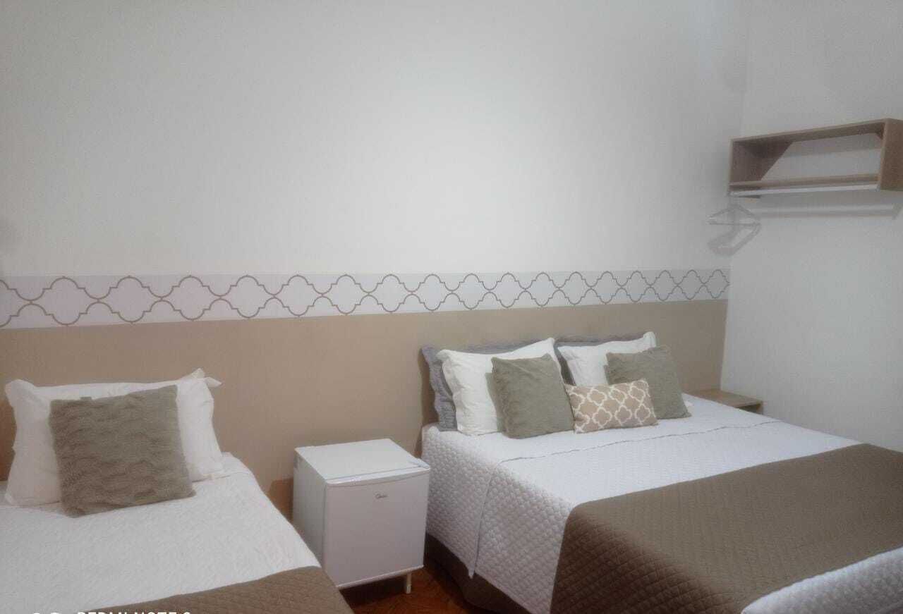 A hotel room with two beds, a small bedside table, a white heater, and a shelf above the bed.