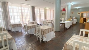 Un comedor con varias mesas y sillas, ventanas amplias con cortinas y piso de baldosas.