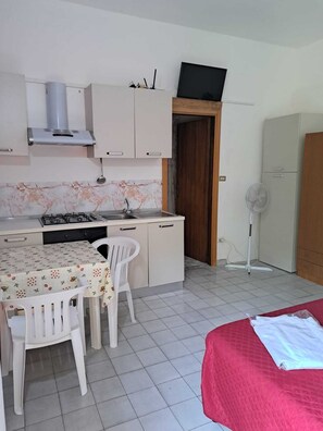 Une cuisine compacte équipée d’une cuisinière à gaz, d’un évier et d’un four à micro-ondes. On y trouve une table à manger avec des chaises et un lit recouvert d’une couverture rouge.