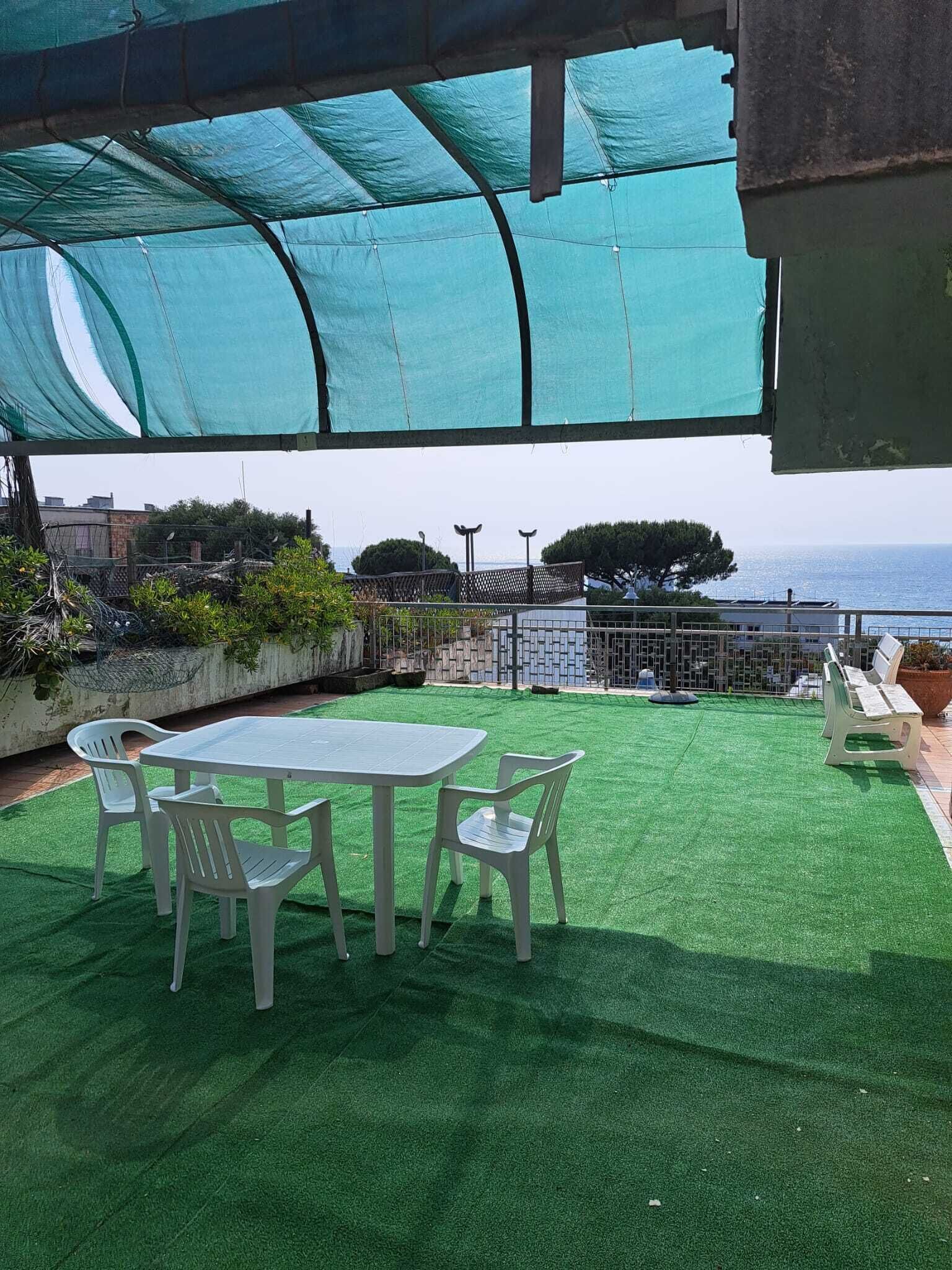 Um pátio externo com mesa e cadeiras, um gramado sintético verde e vista para o mar.