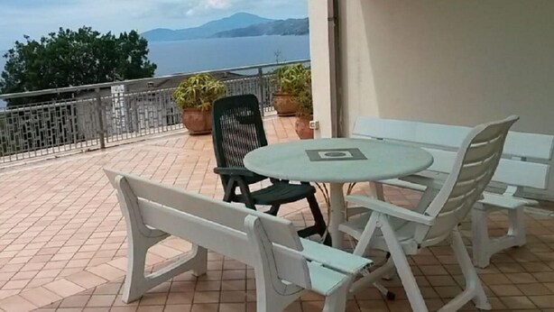 A terrace with a round table and four chairs, a potted plant, and a view of the sea.