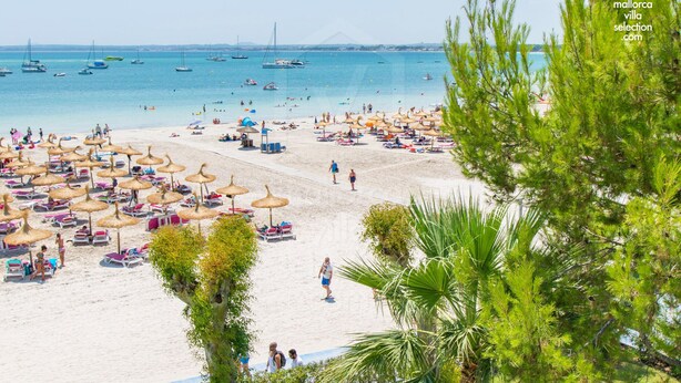 Ein Strand mit vielen Sonnenschirmen und Liegestühlen, Booten im Wasser und wolkenlosem Himmel.