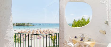 A balcony with a view of a beach and sea, a table with chairs, and a small window.