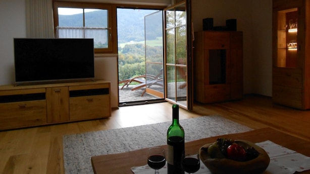 A living room with a television, a wooden cabinet, a glass table with a bottle and glasses, and a large window with a view of mountains. - Spacious Schneiderhof vacation apartment with terrace (Neukirchen)