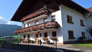 A two-story building with balconies adorned with flowers, a stone wall in front, and a mountainous backdrop. - Spacious Schneiderhof vacation apartment with terrace (Neukirchen)