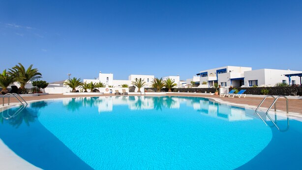 A swimming pool with clear blue water, surrounded by white buildings and palm trees. - Spacious Apartment "Casa Nancy" with Mountain View, Pool, Wi-Fi & Terrace (Yaiza)