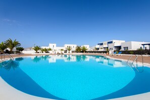 A swimming pool with clear blue water, surrounded by white buildings and palm trees.
