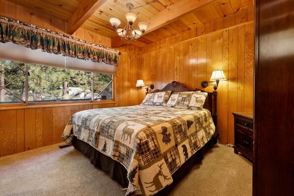 Une chambre à coucher dans un chalet en bois, avec un grand lit, deux lampes de chevet, une fenêtre donnant sur les arbres et un ventilateur de plafond.