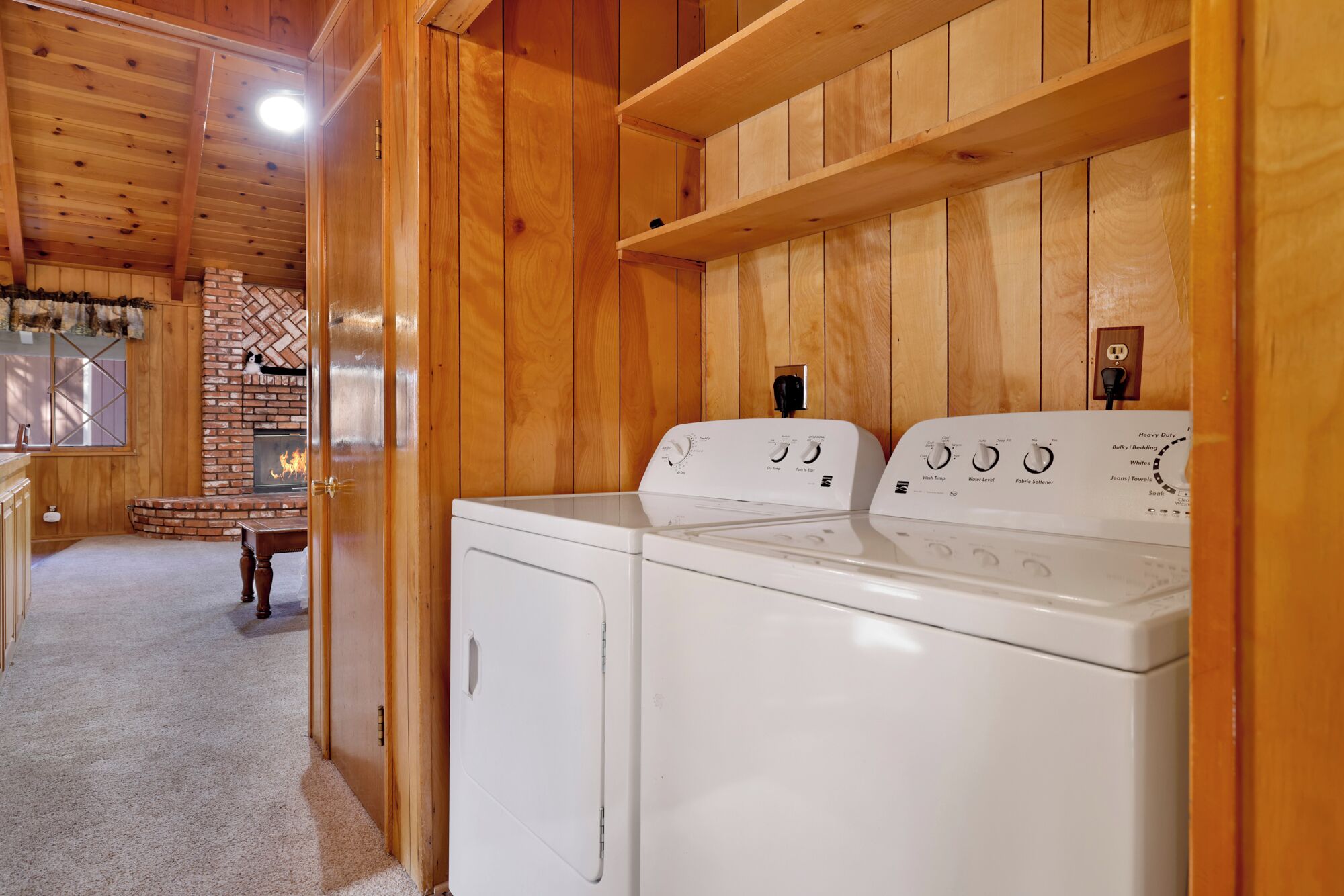 A laundry room with two white washers and dryers, wooden walls, a brick fireplace, and a window with blinds.