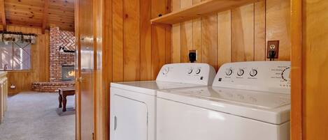 A laundry room with two white washers and dryers, wooden walls, a brick fireplace, and a window with blinds.
