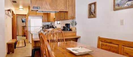 A wooden dining table with chairs, a kitchen area with wooden cabinets, a clock, and a painting on the wall.