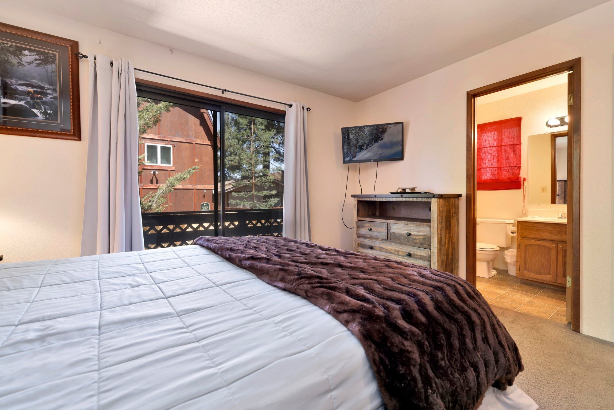 Ein Schlafzimmer mit Bett, einem fensterbeprägten Fenster, einem Fernseher und einem durch eine offene Tür einsehbaren Badezimmer.
