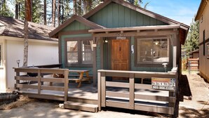 Une petite cabane avec une terrasse en bois, une pancarte d’accueil et une autre faisant la publicité du site BigBearVacations.com.