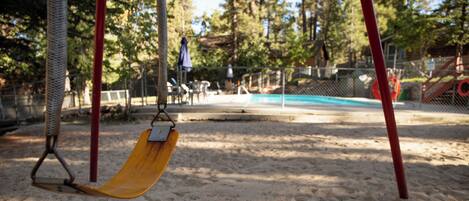 Un parque infantil con arena, una resbaladilla y una piscina cercada al fondo.