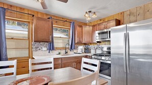 A modern kitchen with wooden cabinets, stainless steel appliances, and a dining area with white chairs and a wooden table. - Lakeside 109 (Big Bear Lake)